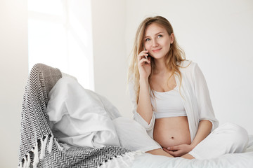 Pregnant woman talking to doctor on phone, anticipating her future healthy baby smiling in her bedroom