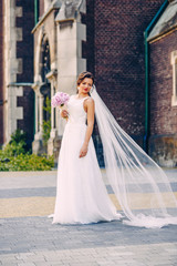 Obraz premium Stunning beautiful bride with the bouquet in a white dress standing alone outdoors on the background of old historical building