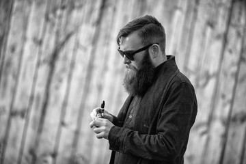 Vape man. E-liquid filling process. Outdoor portrait of a young brutal white guy with large beard vaping an electronic cigarette opposite the old wooden fence in the village. Black and white photo.