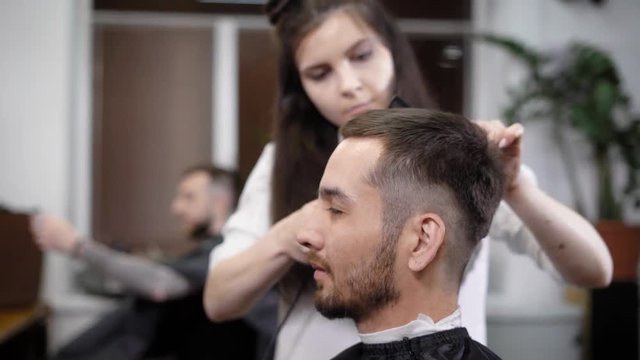 An adult and stylish woman who is a young and practicing stylist makes a new immigration for a beauty salon, a man with a beard expects a haircut