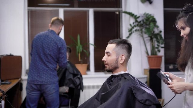A stylish and young woman hairdresser takes a hair trimmer in her hands and begins to make hair styling and haircut for a beauty salon for a man