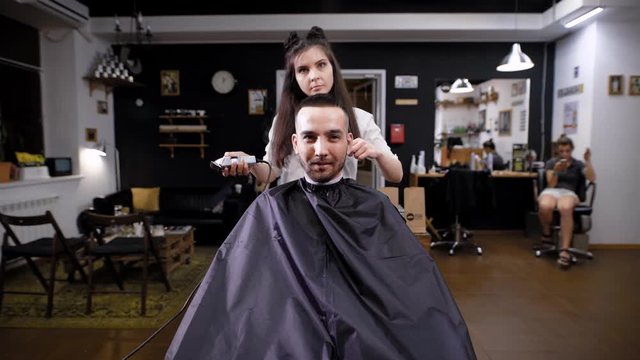 A pretty woman, a hair stylist, shortens the length of her hair with trimmra for her hair, a man who sits in an armchair awaits the completion of the image