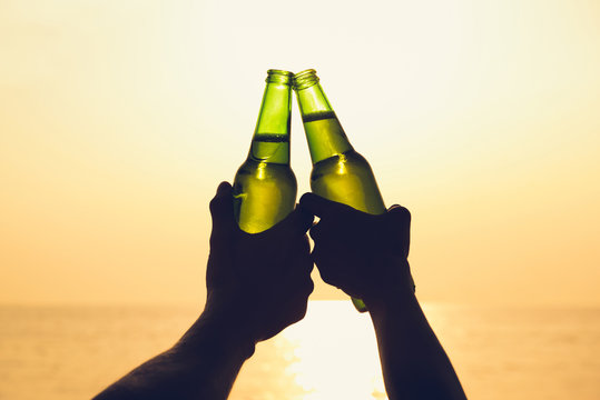 Couple hands holding beer bottles, clanging at the beach in summer sunset