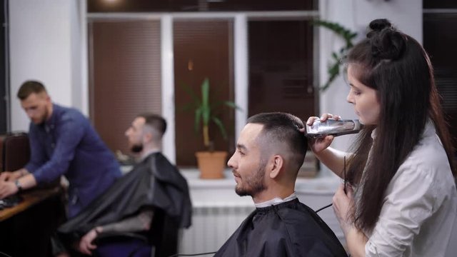 A woman with long dark hair who turns out to be a hairdresser, cuts the hair of a client of barbershop, a man wants to change the immige of his hairstyle