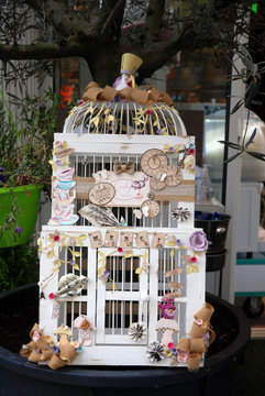 Marché de Fréjus : Cage à oiseau blanche ornée de petits paniers, de fleurs, de magnets et de feuilles pastelles 
