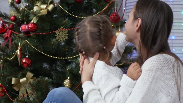 Cute little girl having fun and kissing her mom next to Christmas tree