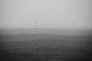 Black and white cityscape or view of a city downtown from a distance with many layers of forests in the middle showing sun rays shot at dusk from a mountain