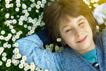 Naklejka premium Handsome happy smiling young boy lying on summer meadow green grass with daisy. Attractive smiling teen boy posing over nature background, image toned.