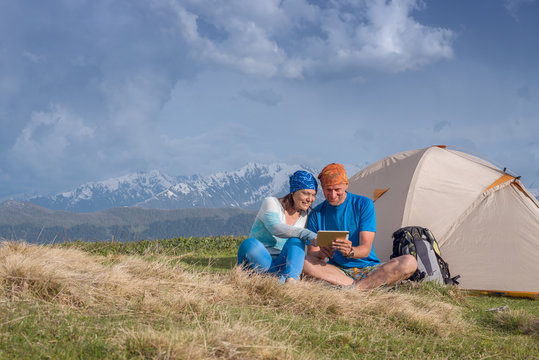 Couple of happy travelers sit next to tent and use tablet