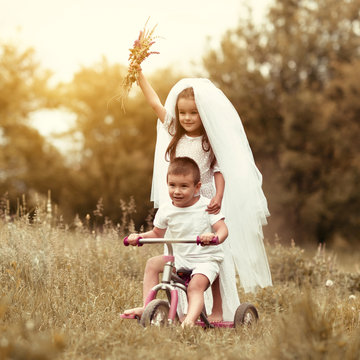 Young bride and groom playing wedding summer outdoor. Children like newlyweds on bicycle. Little girl in bride white dress and bridal veil walking with her little boy groom. Bridal, wedding concept