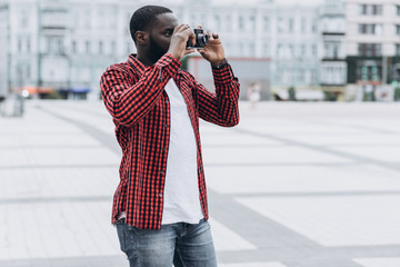 Outdoor summer smiling lifestyle portrait of handsome and happy Afro American tourist having fun in the city in Europe with camera travel photo of photographer making pictures in hipster style