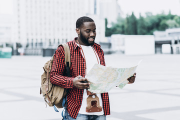 Portrait of handsome Afro American tourist wearing casual clothes with old camera and map in modern city. He is looking at camera and smiling