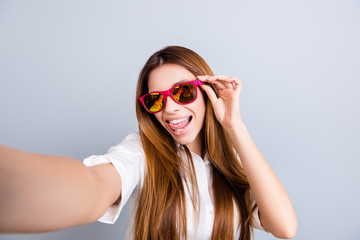 Selfie mania! Funky mood. Attractive young lady is making a selfie on the camera, flirty and playful. In pink trendy sunglasses, with tongue out, on pure light grey background