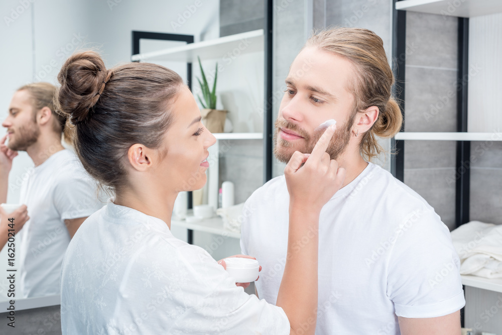 Wall mural Smiling young woman applying face cream to bearded husband in bathroom