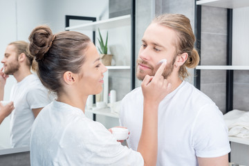Smiling young woman applying face cream to bearded husband in bathroom