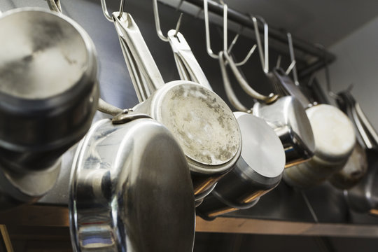 Close up of stainless steel pots and pans hanging on metal hooks on a shelf in a restaurant kitchen.