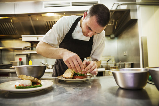 Chef standing in kitchen, wearing apron, plating food.