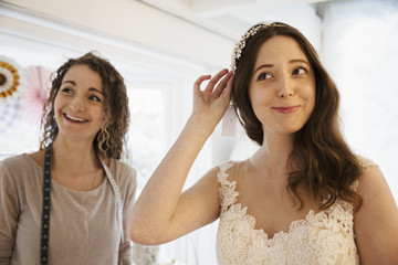 A young woman trying on hair accessories, and veil with a white lace overlay wedding dress, assisted by a retail advisor. 