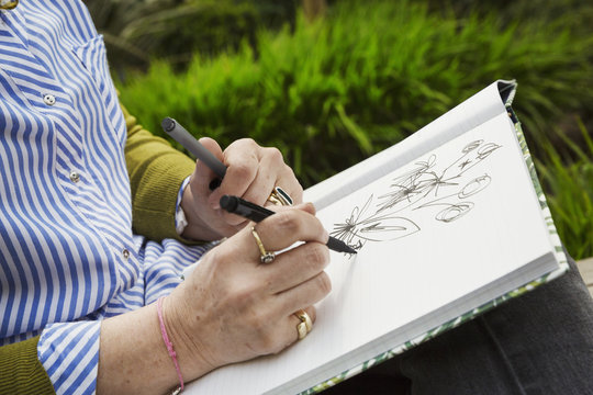 Close up of a woman sitting in a garden, drawing flowers in a sketchbook.