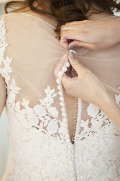 Hands doing up the small buttons on the back of a wedding dress with a net bodice and lace detail.  A bride getting ready. 