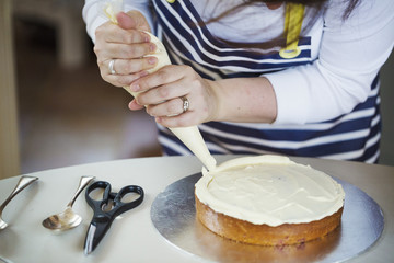 Close up of person wearing a blue and white stripy apron, holding a piping bag, decorating a cake with cream, spoons and scissors on table.