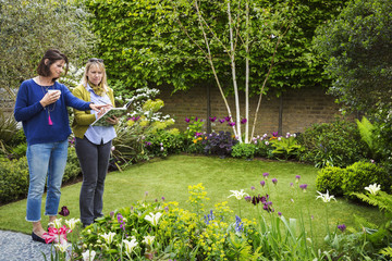 Two women standing in a garden on a lawn surrounded by flowerbeds, discussing garden design.