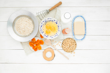 Raw eggs in a bowl, butter, milk bottle, peanuts, whisk, spoon, bowl of cereal, dried fruits on a light wooden table against a white background. The ingredients for the cookies.