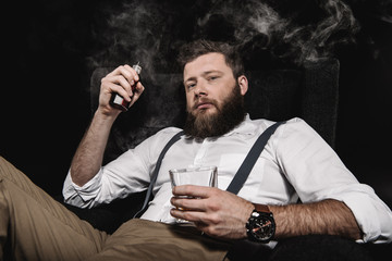 stylish man in white shirt and suspenders vaping and holding whisky glass, isolated on black