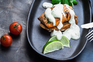 Fish steak with white sauce in a skillet with basil close-up, fork and knife on a dark metallic background. Fish meal.