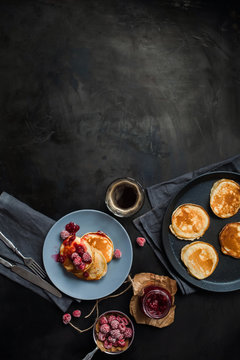 Pancakes with raspberry jam on a blue plate, frozen raspberries, a jar of raspberry jam, a cup of coffee, a frying pan with pancakes, fork and knife on dark metal table top view. Breakfast.