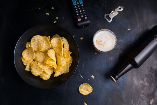 Plate of chips, remote control TV, beer, bottle opener top view of a dark metal table.