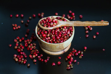 Pink pepper peppercorns in wood bowl and spoon on black background.  Copy space. Selective focus.