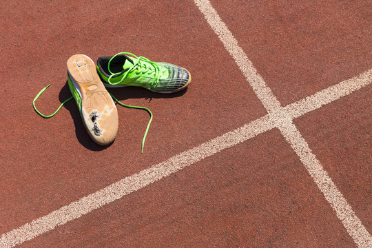 A pair of broken green running shoes with big holes in the sole laying on a running track