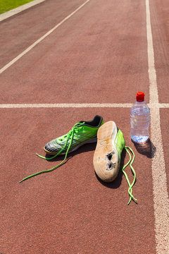 A pair of broken green running shoes with big holes in the sole laying on a running track besides a water bottle.