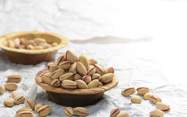 In the front there is brown wooden bowl of pistachio place and a another in the background with some pistachio fallen around.