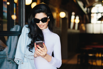 Horizontal portrait of magnificent brunette woman in white clothes and sunglasses sitting in cafe using her smartphone messaging with friends using free internet conncection. Beauty and youth concept