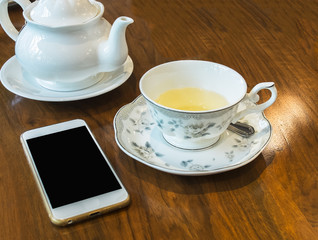 Antique Tea Set with White Jar and Beautiful Cup Full of Herbal Tea on Wooden Table for Tea Break Time or Breakfast and Blank Screen of Mobil Phone for Mock up Photo or input Text. Business Concept