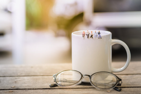 Group of miniature figures sitting and read a book, read newspaper, waiting, talking and relax on white cup of hot coffee with reading glasses on wooden table