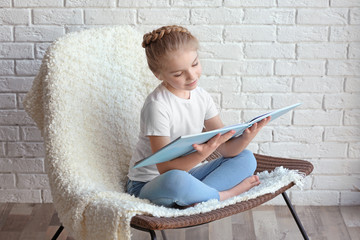 Small girl sitting on chair with book in light room