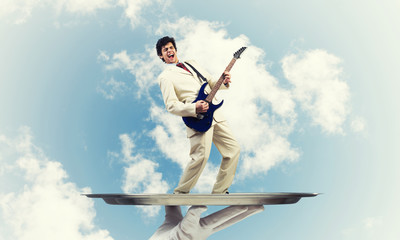 Businessman on metal tray playing electric guitar against blue sky background