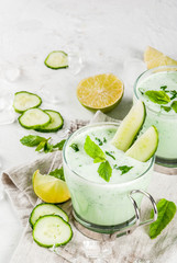 Summer food. Refreshing dishes. Cold soup of cucumber, avocado, with herbs and mint. With serving glasses, with slices of cucumber. On a white concrete table, with ingredients. Copy space