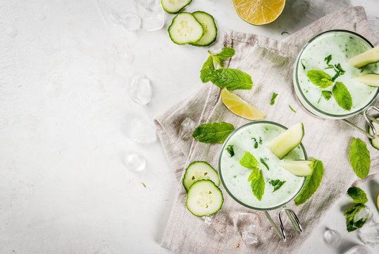 Summer food. Refreshing dishes. Cold soup of cucumber, avocado, with herbs and mint. With serving glasses, with slices of cucumber. On a white concrete table, with ingredients. Copy space
