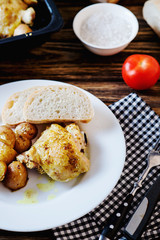 Hearty and delicious dinner, baked chicken legs and thighs with potatoes, onions, tomatoes, spices, herbs and white bread on a dark wooden background