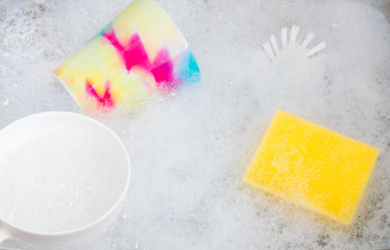 Colorful dirty mug, white cup, sponge and a brush soaking in foamy soap water in the kitchen sink.