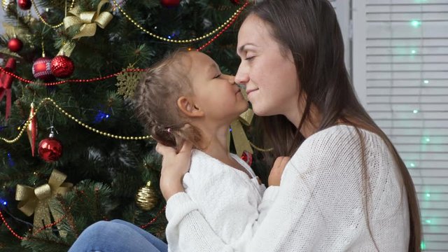 Cute little girl having fun and kissing her mom next to Christmas tree