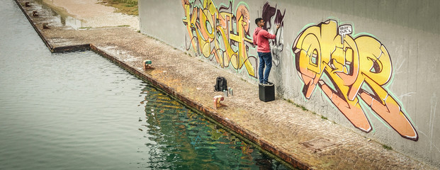 Young graffiti writer painting with color spray his picture on the wall - Top with of contemporary artists at work - Urban lifestyle,street illegal art concept - Focus on man face - Contrast filter