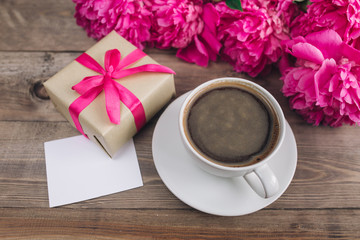 A cup of coffee , pink peonies pattern and gift box and paper card on wooden background . Good morning