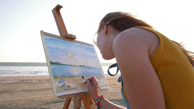 Young woman artist painting landscape in the open air on the beach, slow motion