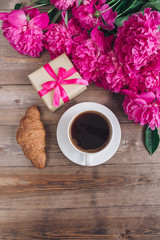 A cup of coffee , pink peonies pattern and gift box and croissant on wooden background . Good morning