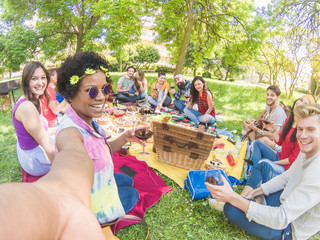 Fototapeta premium Happy afro woman taking selfie at picnic outdoor with her friends - Young students having fun while eating bbq lunch and toasting red wine - Youth concept - Focus on afro girl - Summer color tones